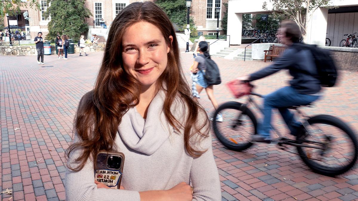 Keegan Lee posing for a portrait on brick pathways on the campus of UNC-Chapel Hill. She's holding her phone, which has a sticker on it reading: 